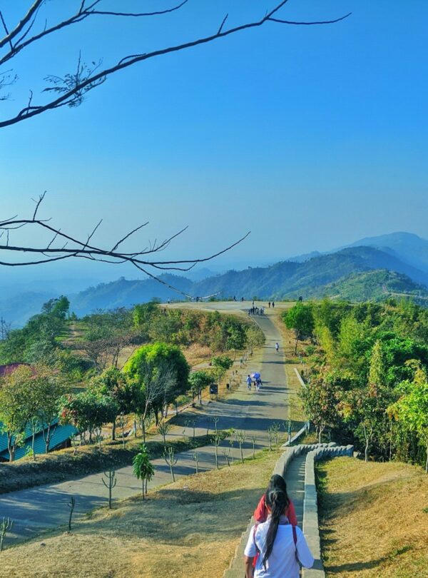 woman in white shirt walking on pathway near green trees and mountain during daytime